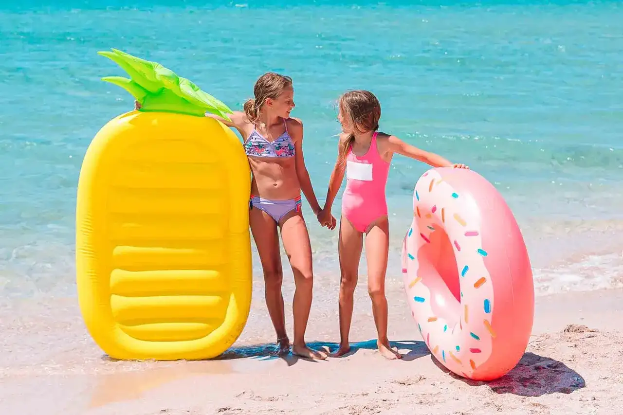 Dwie dziewczynki na plaży z materacami do pływania: żółtym ananasem i różowym pączkiem.